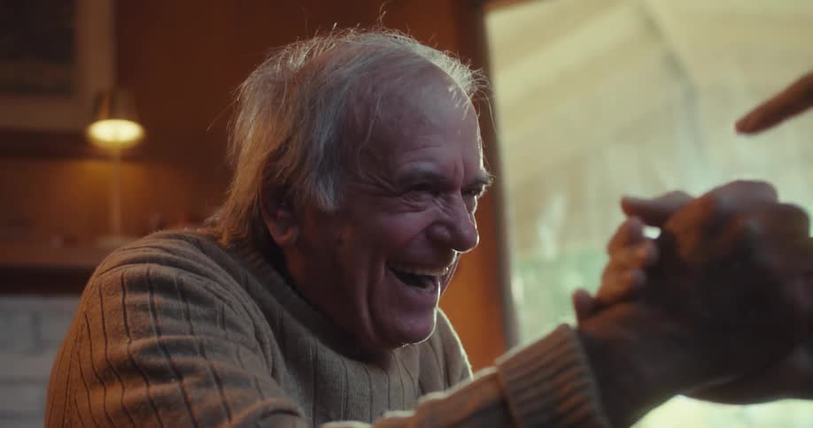 A smiling older man with white hair engages in a handshake with another person their hands tightly clasped