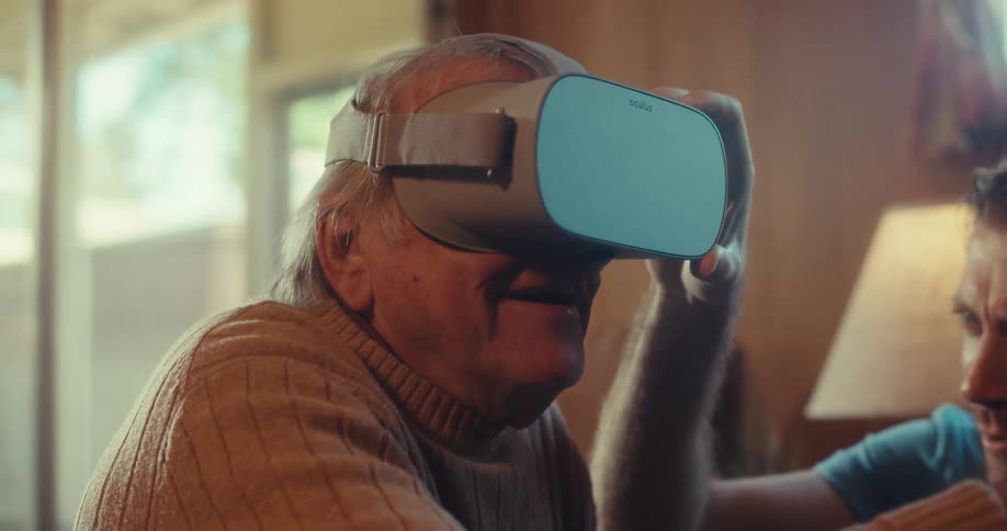 An older man is using a virtual reality headset while a younger man helps him