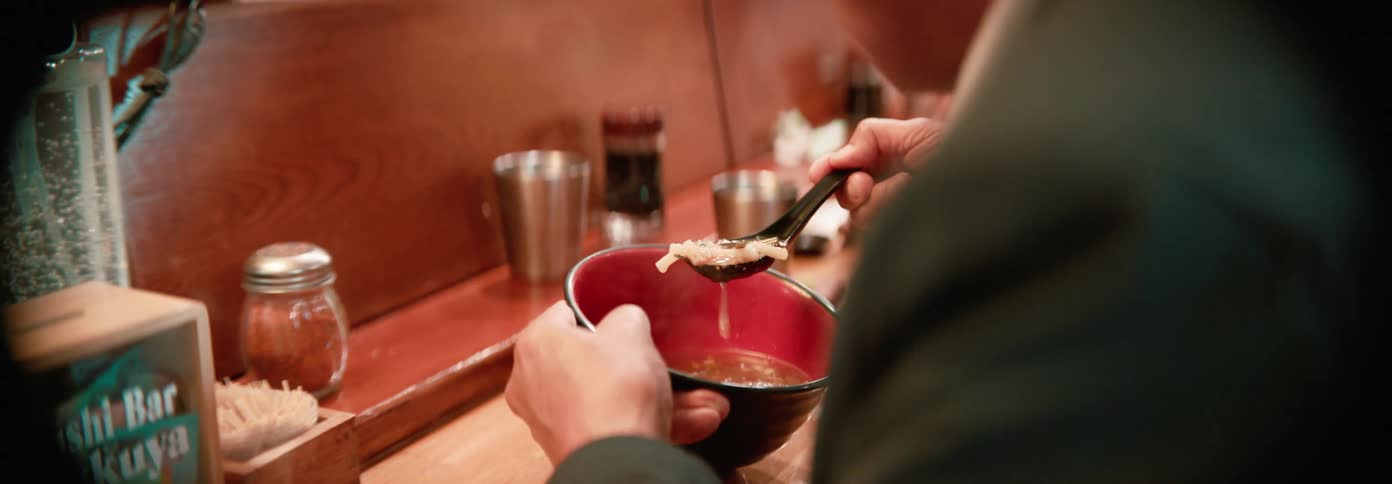 A man drinks soup from a red bowl at a sushi bar counter