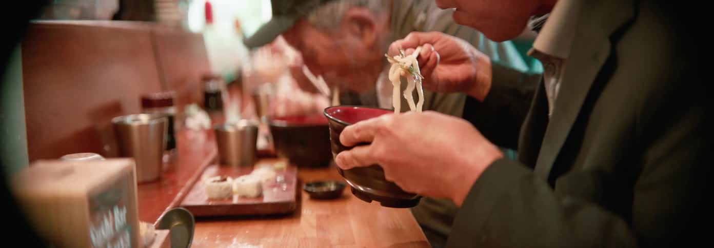 A group of people are eating noodles from bowls with chopsticks at a wooden counter in a dimly lit setting