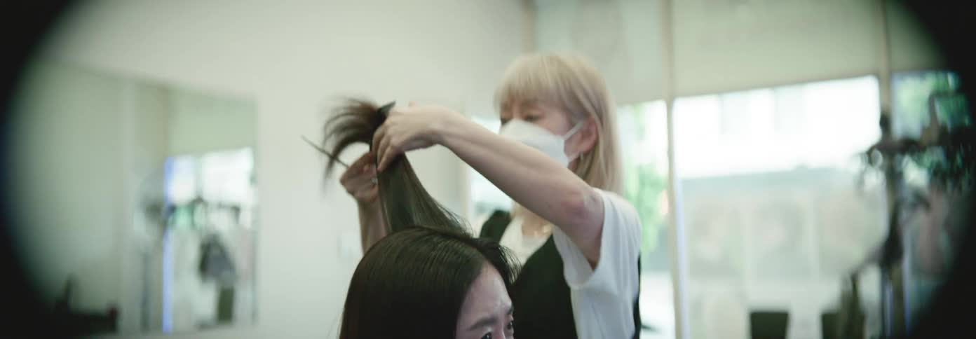 A woman with long dark hair sits in a salon chair while a masked hairstylist cuts her hair with scissors