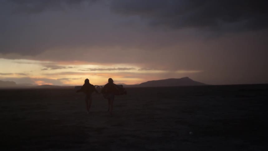 Two people run across the ground toward the setting sun