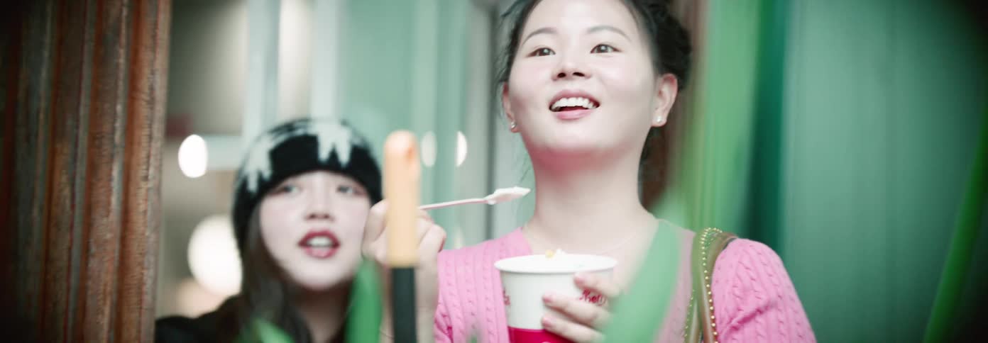 A young woman smiles while eating from a small cup with a spoon as another woman wearing a patterned beanie stands nearby