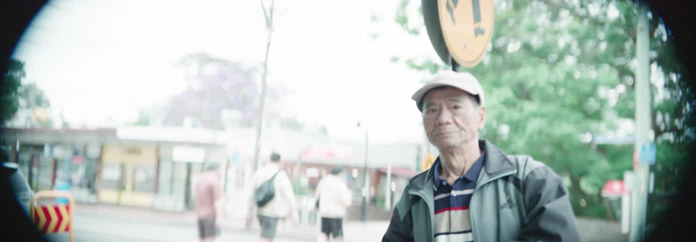 An elderly man waves with a gentle smile while standing on a city street corner