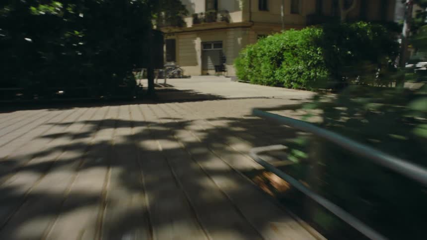 Fast motion ground view zooming toward a courtyard with benches, green space, buildings, pedestrians, and people riding by on bikes.