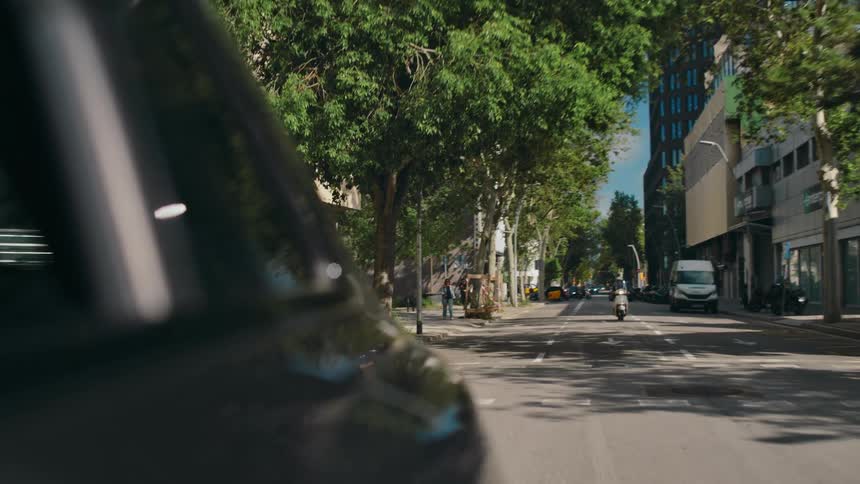 POV from the side of a moving car looking back toward the tree-lined city street behind as a motorcyclist approaches and drives past when the car turns.
