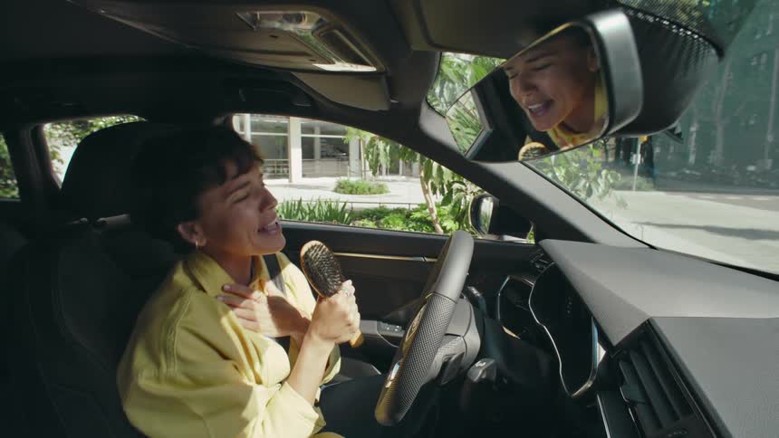A woman driving a car with her reflection in the rearview mirror sings into a hairbrush she is pretending is a microphone as she happily sings and sways to music.