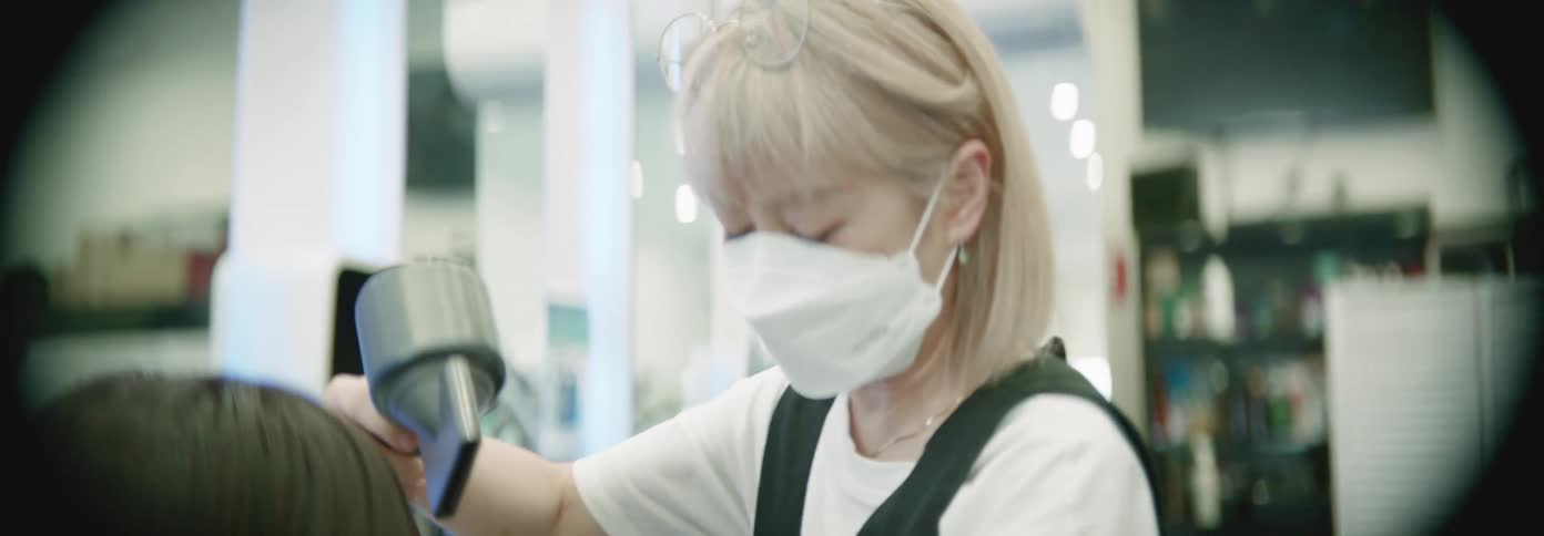A hairstylist wearing a face mask is blowdrying a clients hair in a salon