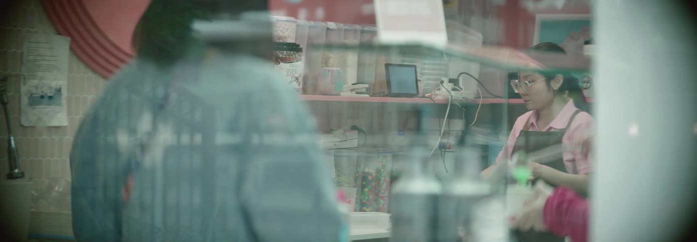 A young woman in an apron prepares food containers behind a counter seen through a glass window