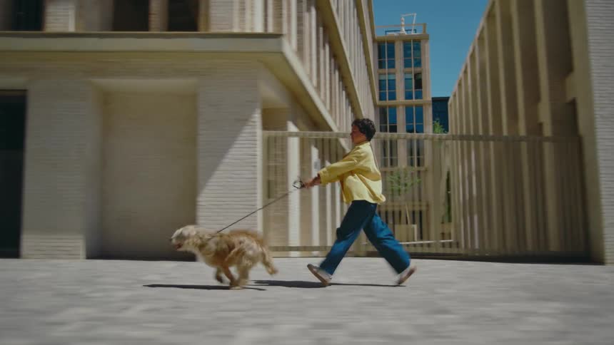 A woman struggles to keep up with her dog on a leash as he runs excitedly pulling her along past buildings on a city sidewalk