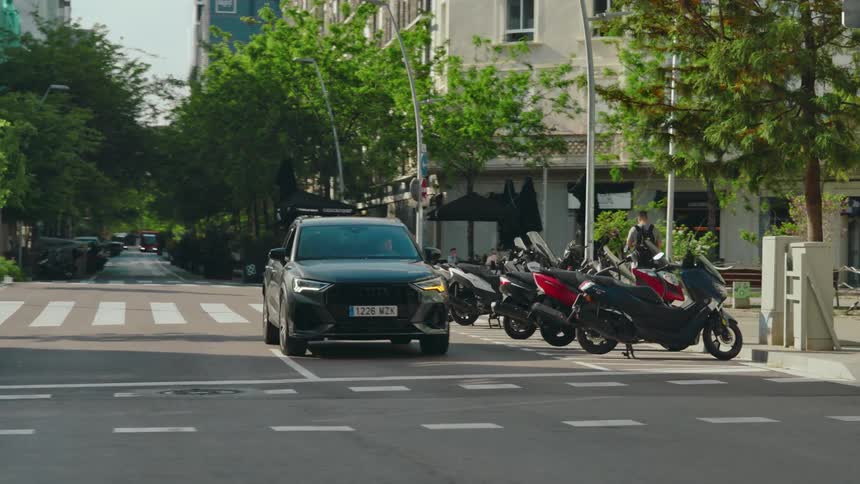 A person drives a vehicle down a tree-lined city street and across a zebra crossing and past a row of moped scooters, then turns on the blinker and turns left at the junction.