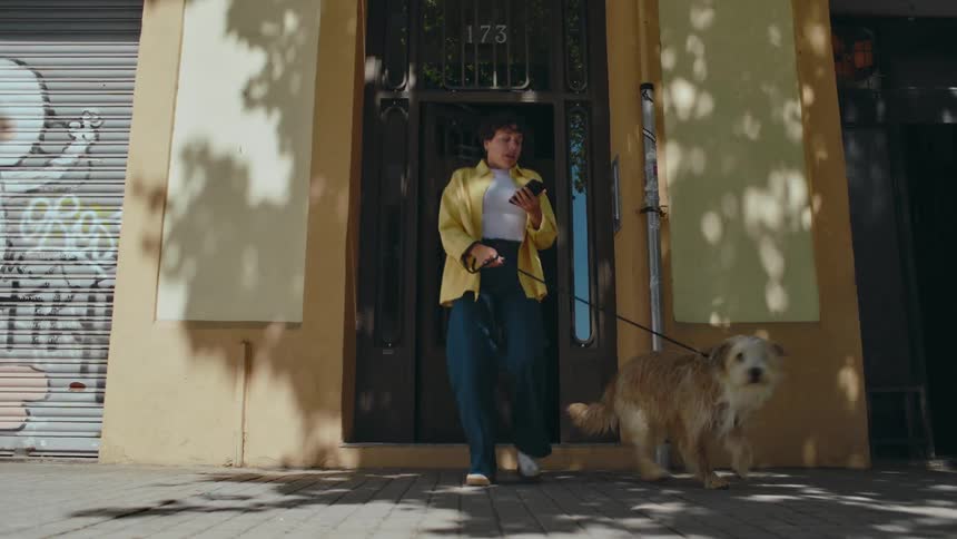Woman walking out the door of a building in the city with her dog on a leash while looking down at her phone as the dog looks around and then excitedly runs to the right yanking her along with him.