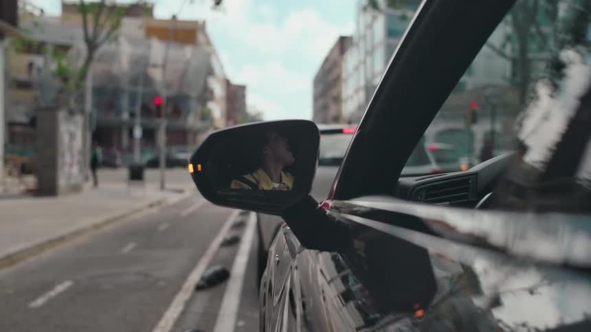 The side mirror of a dark car reflects a woman singing and dancing inside her vehicle as she is stopped at a traffic light along a busy city street and is passed by several people on bicycles and pedestrians.