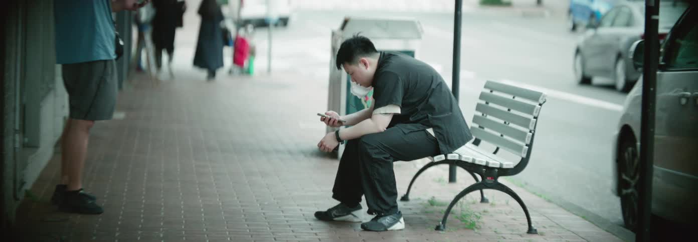 A man dressed in dark scrubs is sitting alone on a bench at a bus stop focused on his phone