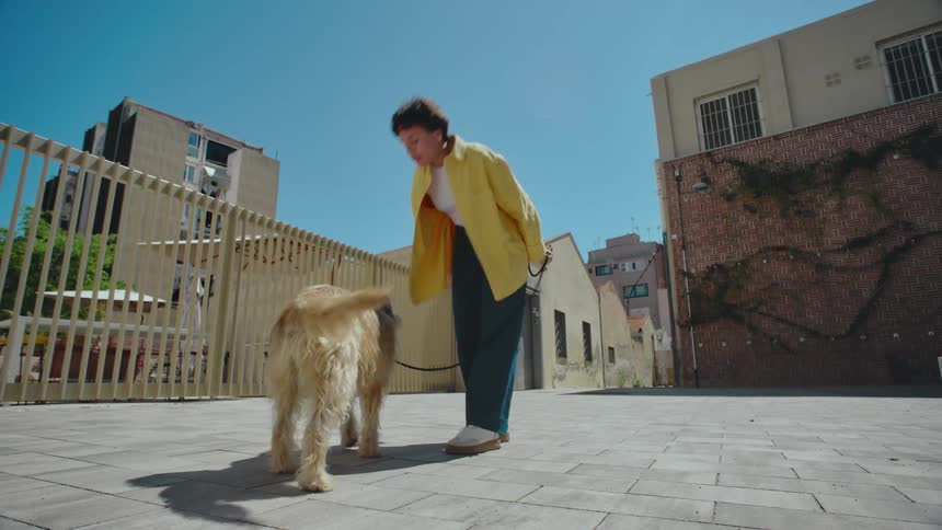 A woman trains her dog with a treat in a sunlit urban courtyard