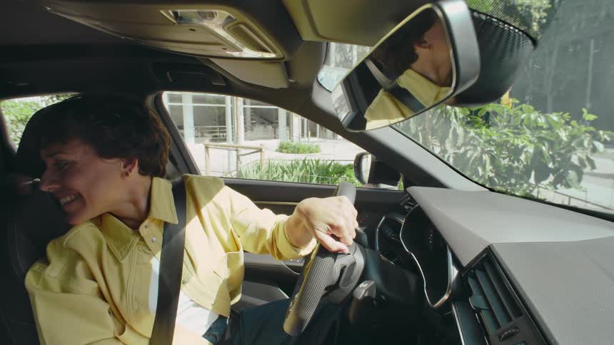 A woman in the driver's seat of a car smiles as she adjusts her rear view mirror, and then turns to engage with someone in the backseat.