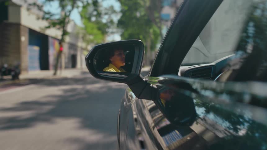 POV from the outside of a moving car looking at the driver's side mirror that is reflecting an excited female driving along a city street during the day
