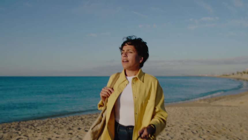 A joyful woman walks her dog along a sunny beach as she looks around searchingly, and then waves excitedly to someone in the distance.