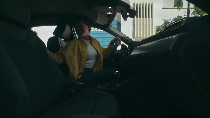 A woman in a yellow jacket enters the driver's seat of a car, then fastens her seatbelt while smiling and talking to someone in the back.