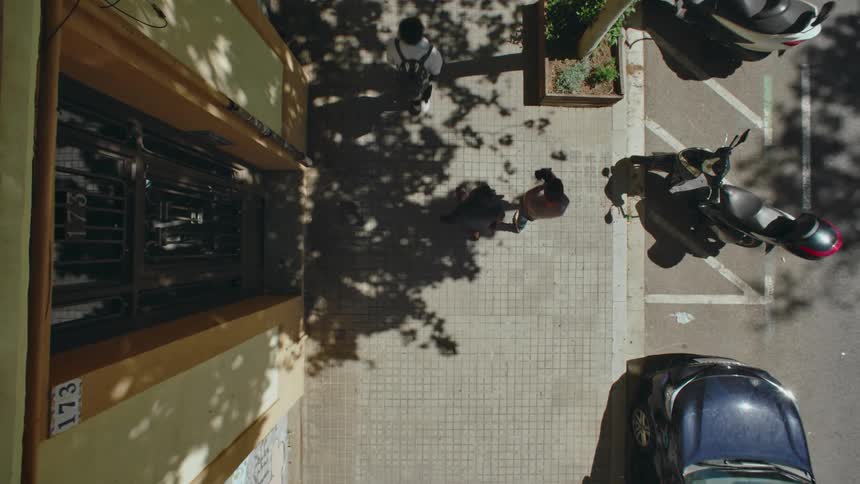 Three people are walking on a sunlit city sidewalk captured from a topdown aerial view