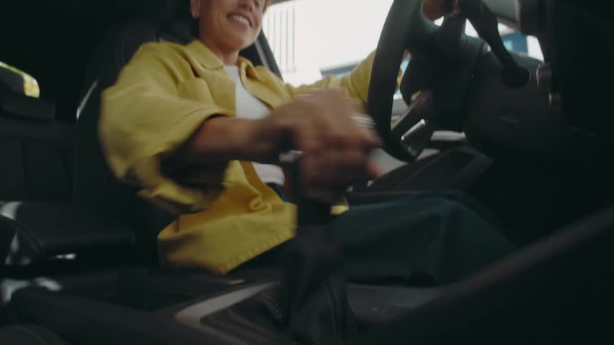 View looking up at a smiling woman with short curly dark hair wearing a yellow jacket as she sits in the driver's seat of a car shifting gears