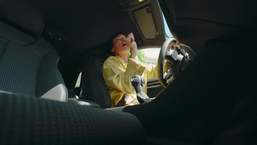 View from the car floorboard below as a woman happily pretends to sing karaoke with a hairbrush while sitting in the driver's seat waiting at a stoplight.