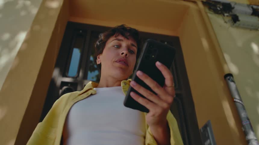 Close up view panning down of a woman with short dark curly hair standing outside the front door of a building looking down at a black smartphone in her hands and heavy exhaling before suddenly being pulled to the side by a force offscreen.