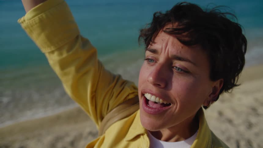 A woman at the beach furrows her brows as she looks for someone in the distance, and then breaks into a big smile and starts waving when she see them.