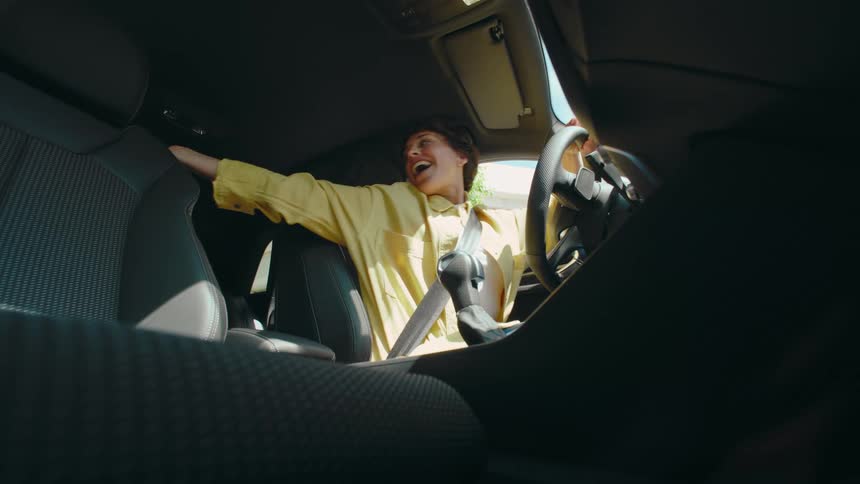 POV from the passenger floorboard of a woman in a driver's seat of a car as she chats excitedly with someone in the back seat before starting to drive