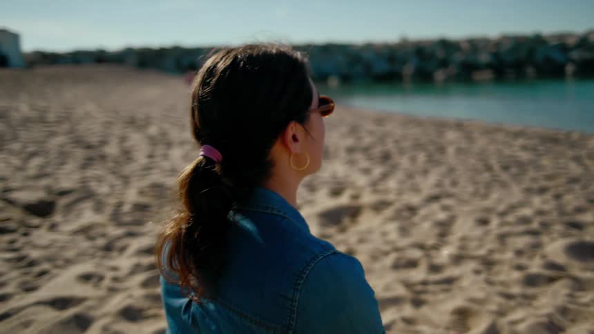 Two women sit on a sandy beach looking toward the crystal blue water and chatting happily as one of them throws a ball for her dog to fetch.