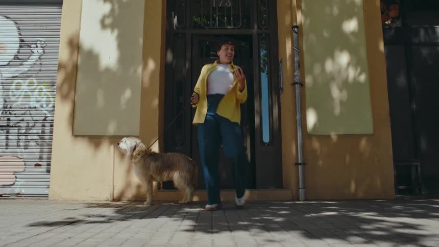 A woman steps outside with her dog on a leash and looks at her phone when the dog suddenly pulls and starts running, dragging her along.