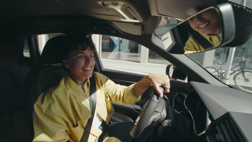 A woman wearing a yellow shirt is driving a car and excitedly talking to a passenger in the back seat. After coming to a stop, she picks up a brush and sings into it like a microphone.