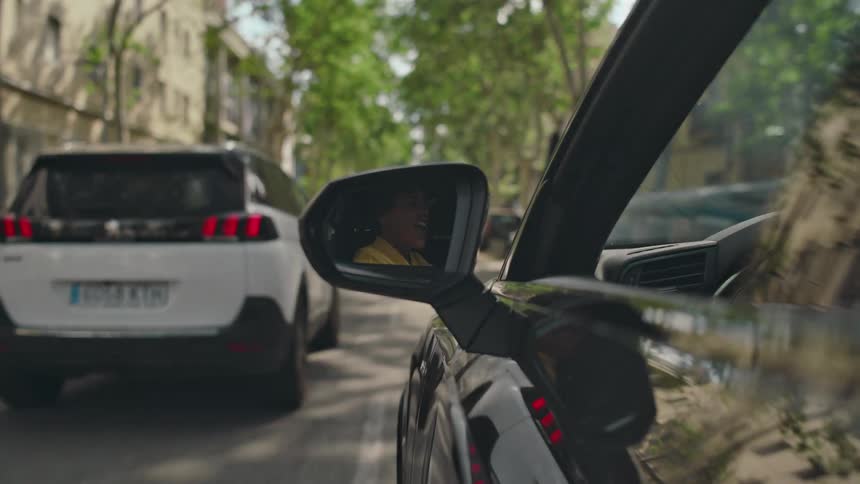 Side mirror reflects a woman singing while she drives along a street during the daytime