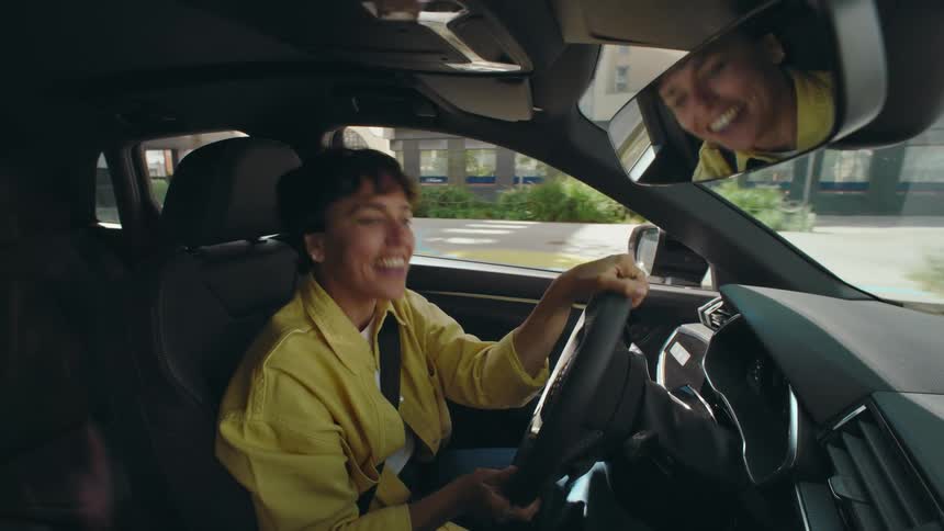 A woman wearing a yellow jacket drives through the city on a sunny day singing with excitement and drumming her hands on her steering wheel as she listens to music.