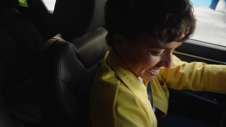 A woman sitting in the driver's seat of a car looks behind her with a big smile to make sure the way is clear and gets ready to back up.