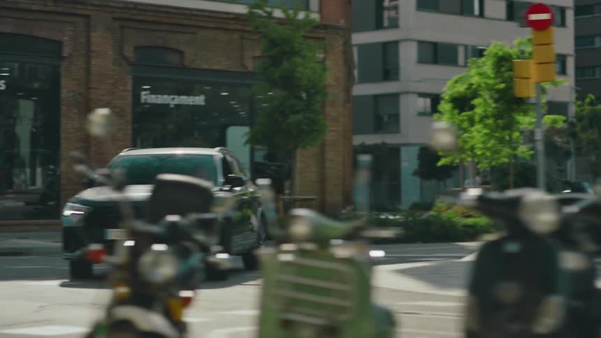 Black SUV driving around a curve at an intersection in a city where multiple scooters and motorcycles are parked and signal lights direct traffic and pedestrians