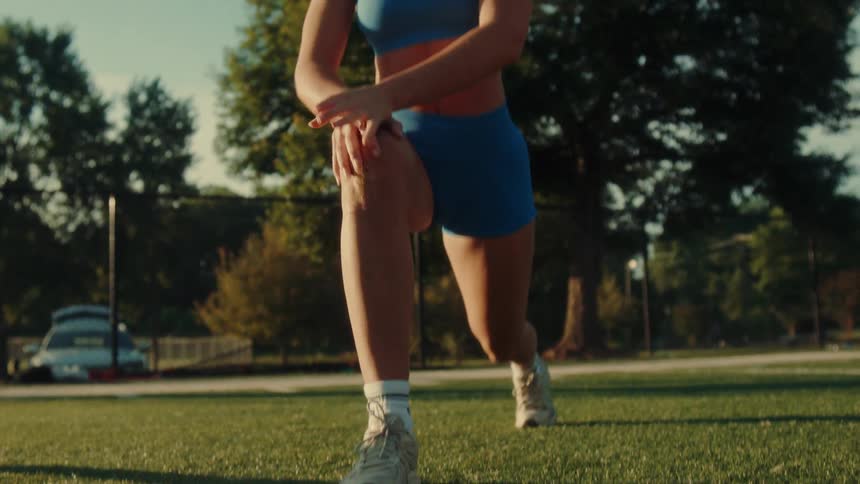 A blonde woman in blue athletic wear stretches her leg forward while kneeling on a green field with trees in the background