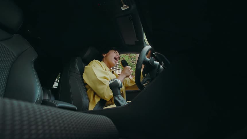 POV from the passenger floorboard of a car panning up and over toward the happy female driver who picks up a brush from the middle console and dramatically pretends to sing into it like a microphone.