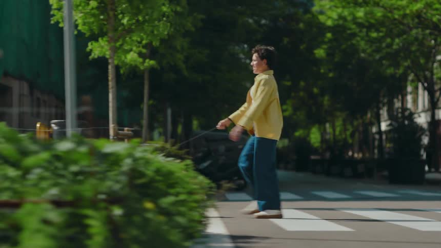 A woman in a yellow shirt and blue pants walks a furry dog across an intersection on a city street lined with trees and buildings