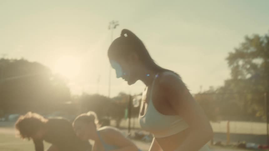 Two women in athletic wear are stretching outdoors on a sports field during golden hour