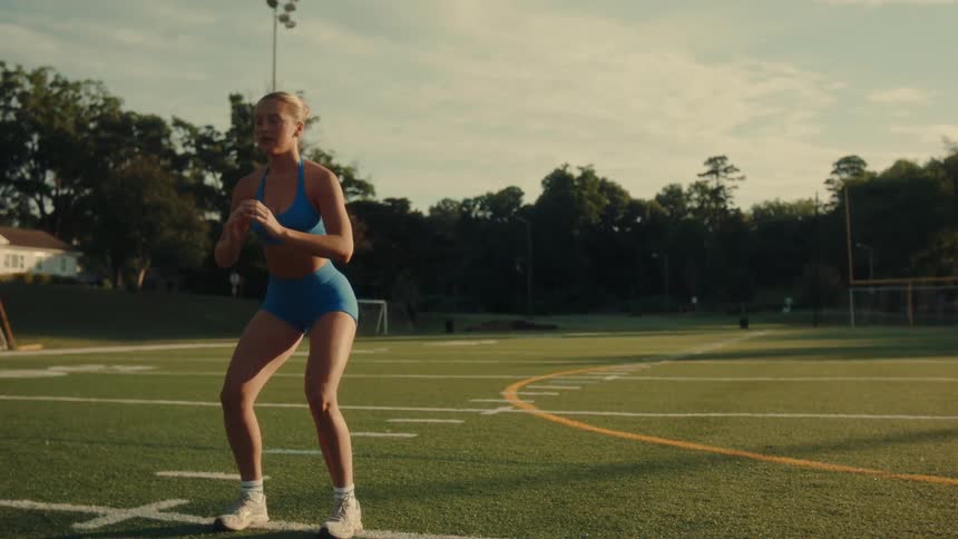 A blonde woman in a blue athletic outfit does squats and lunges on a football field in the early morning light