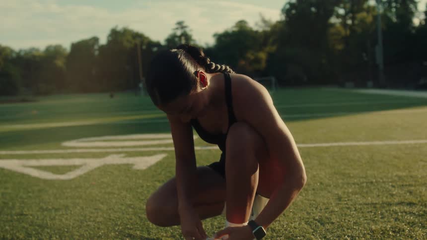 Two women are on a sports field with white lines one standing and the other crouching to tie her white athletic shoe
