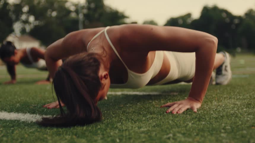 Two women with long dark hair are performing planks on a grassy field under an overcast sky