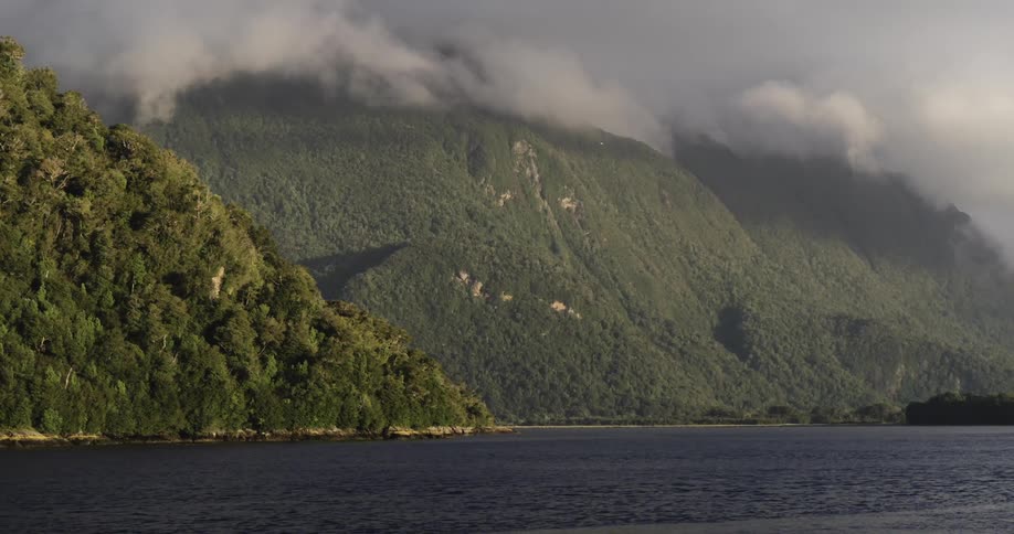 Thick smoke is filling the sky coming from behind some tree covered mountains alongside a lake