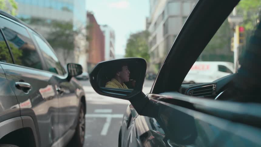 Reflection in side view mirror of a woman driving who is stopped at a stoplight and is singing into her hairbrush as she sways to music and gestures excitedly.