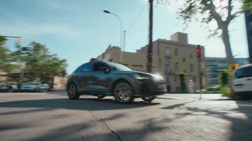 POV from street level as a car drives and turns through a city intersection with buildings, scooters, cars, pedestrians, and a red traffic light in view.
