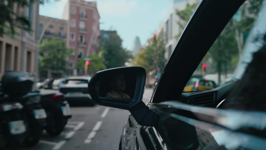 Close up of a vehicle side mirror as it is driven down a street in Barcelona with the Torre Glories skyscraper ahead in the distance.
