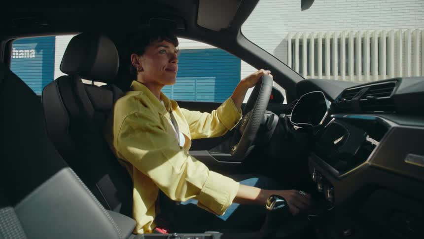 A woman in the driver's seat of a car with her hand on the steering wheel smiles as she adjusts her vehicle rear view mirror, pushes the engine start button, and puts her hand on the stick shift as she looks behind her and prepares to back up.