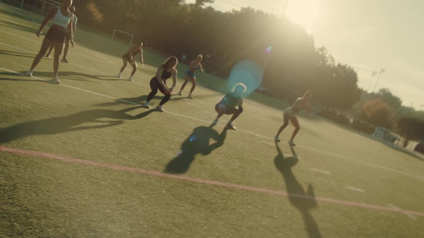 A group of people perform squats together on an outdoor sports field during sunrise or sunset