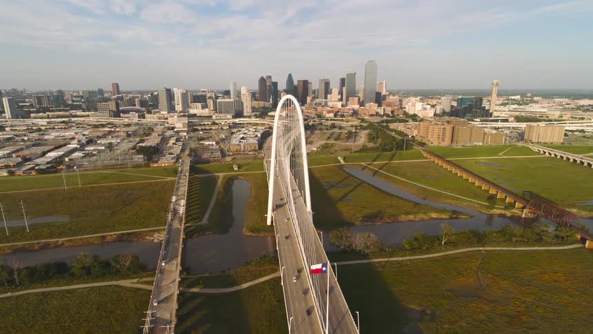 An aerial wide shot captures the iconic margaret hunt hill bridge spanning a river and lush green landscape with the sprawling dallas skyline prominently displayed in the background under a partly cloudy sky